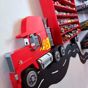 May include: A red wall-mounted display shelf shaped like a semi-truck with a trailer featuring multiple horizontal tiers for toy cars. The truck cab displays a yellow lightning bolt emblem with the number 95, and it is positioned on a black road decal.