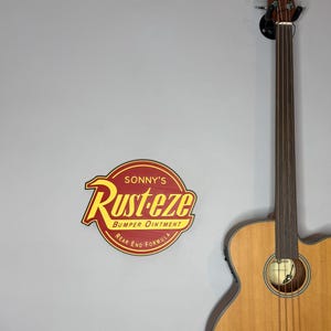 Puede incluir: Un letrero de madera con el texto "Sonny's Rust-eze Bumper Ointment Rear End Formula" en rojo y amarillo. Una guitarra bajo ac&uacute;stica marr&oacute;n est&aacute; montada en la pared a la derecha.