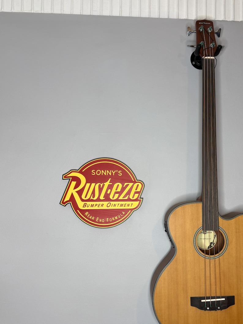 Puede incluir: Un letrero de madera con el texto "Sonny's Rust-eze Bumper Ointment Rear End Formula" en rojo y amarillo. Una guitarra bajo ac&uacute;stica marr&oacute;n est&aacute; montada en la pared a la derecha.