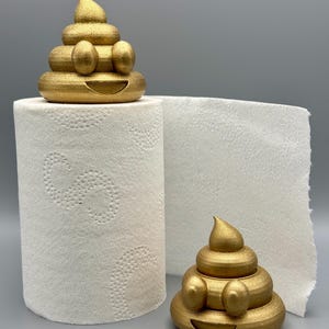 Two gold glittery 3D-printed poop emoji figurines displayed with a roll of toilet paper. One sits on top of the roll, while the other rests beside it on a grey background. Fun bathroom decor idea.