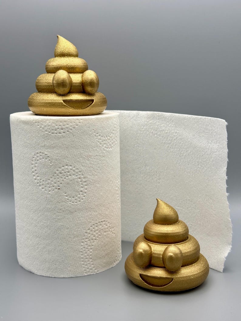 Two gold glittery 3D-printed poop emoji figurines displayed with a roll of toilet paper. One sits on top of the roll, while the other rests beside it on a grey background. Fun bathroom decor idea.