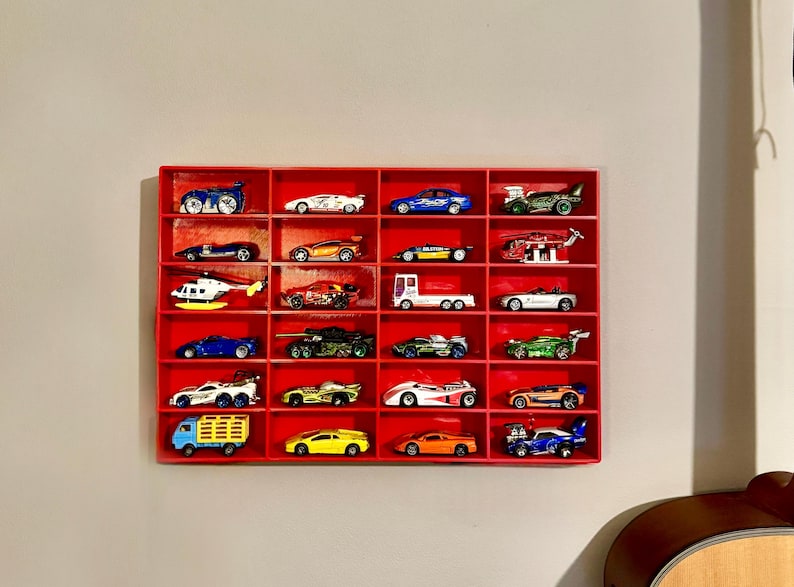 A 3D-printed red wall-mounted display shelf holding 24 toy cars, including sports cars, trucks, and a helicopter. The compact storage solution keeps diecast vehicles neatly organised and easily accessible.