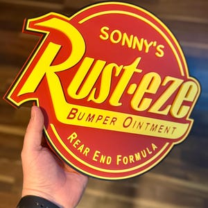 Puede incluir: Un letrero rojo y amarillo con el texto "SONNY'S Rust-eze BUMPER OINTMENT REAR END FORMULA". El letrero es redondo con un borde negro y lo sostiene una persona.