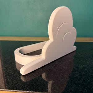 May include: A white, cloud-shaped phone holder. The holder is made of a solid material and has a curved design to cradle a phone. The holder sits on a dark, reflective surface, with a teal background.