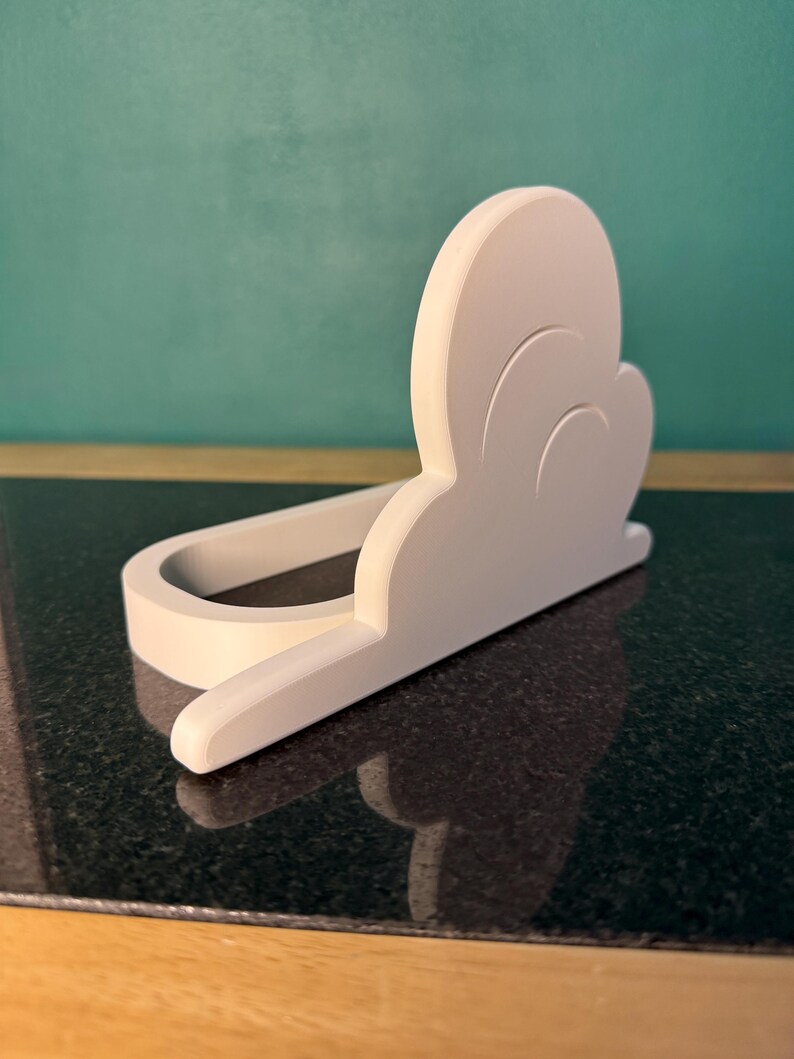 May include: A white, cloud-shaped phone holder. The holder is made of a solid material and has a curved design to cradle a phone. The holder sits on a dark, reflective surface, with a teal background.