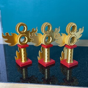 May include: Three gold trophies with a flame design and the number eight. Each trophy has a red base and a cylindrical gold stem. The top features a circular design. The trophies are displayed on a black surface.