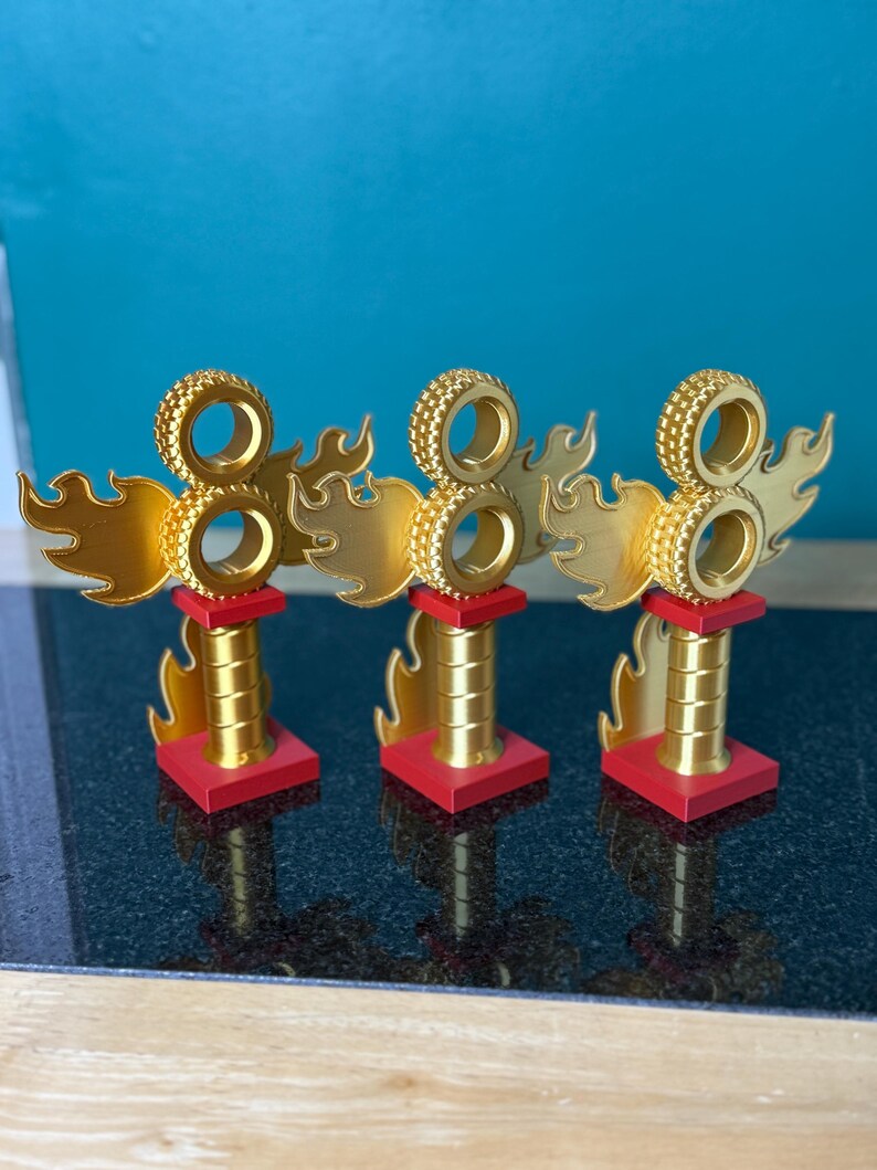 May include: Three gold trophies with a flame design and the number eight. Each trophy has a red base and a cylindrical gold stem. The top features a circular design. The trophies are displayed on a black surface.