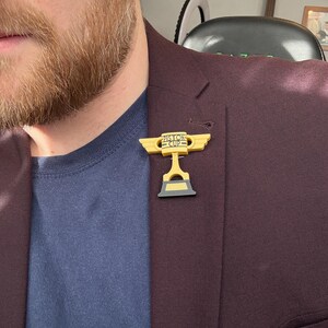 Puede incluir: Un pin dorado y negro del trofeo Piston Cup, con el texto "Piston Cup", est&aacute; prendido a una blazer burdeos. El pin es una r&eacute;plica en miniatura del trofeo de la pel&iacute;cula animada "Cars". La blazer se lleva sobre una camisa azul marino.