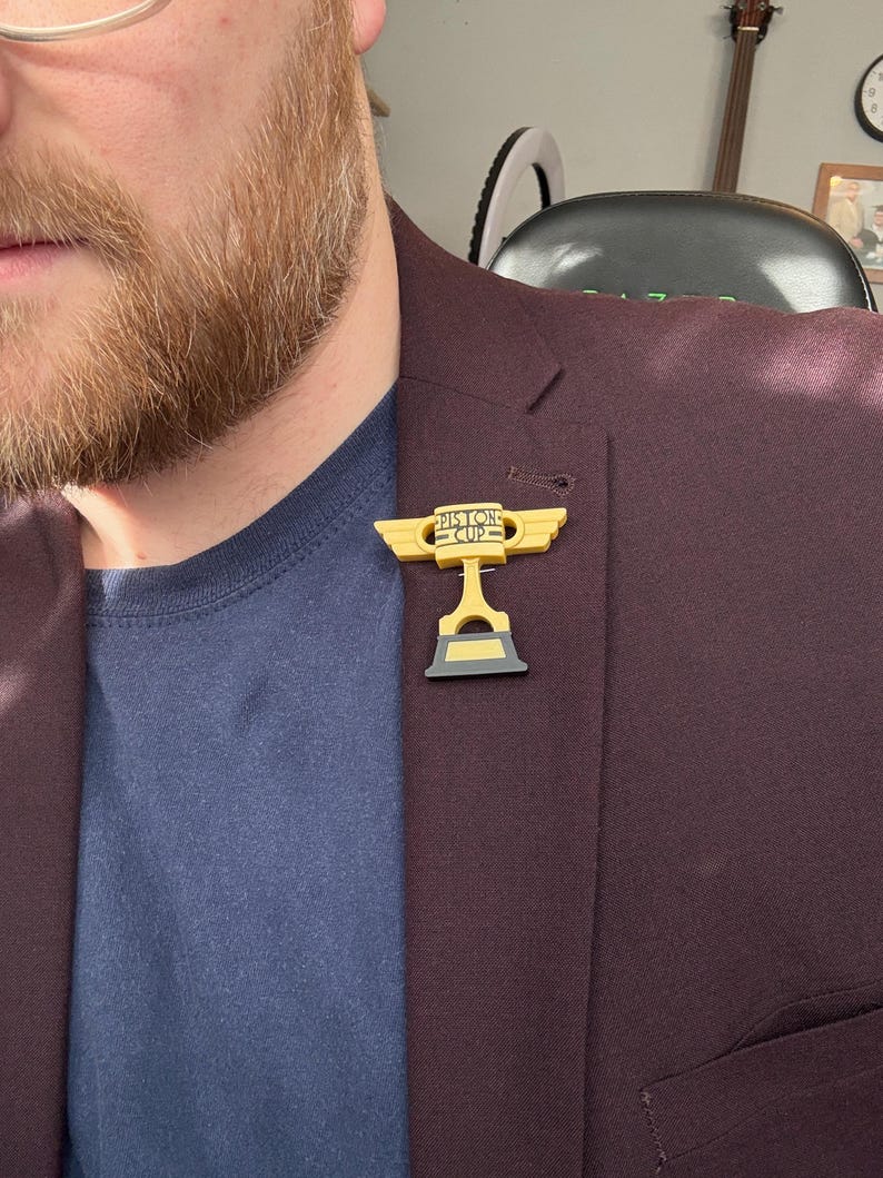 Puede incluir: Un pin dorado y negro del trofeo Piston Cup, con el texto "Piston Cup", est&aacute; prendido a una blazer burdeos. El pin es una r&eacute;plica en miniatura del trofeo de la pel&iacute;cula animada "Cars". La blazer se lleva sobre una camisa azul marino.