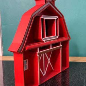 Puede incluir: Modelo de granero rojo con ribete marr&oacute;n oscuro. El granero presenta una peque&ntilde;a ventana, una gran puerta con detalles blancos y una peque&ntilde;a c&uacute;pula en el tejado. El modelo est&aacute; hecho de un material s&oacute;lido y est&aacute; dise&ntilde;ado para mantenerse en pie.