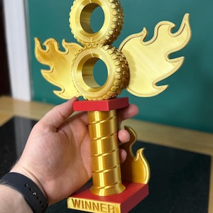 May include: A gold trophy featuring two stacked tyre-shaped elements, flame-shaped wings, and a red base. The trophy has a spiralled column and the word "WINNER" on the base. The trophy is held by a person.
