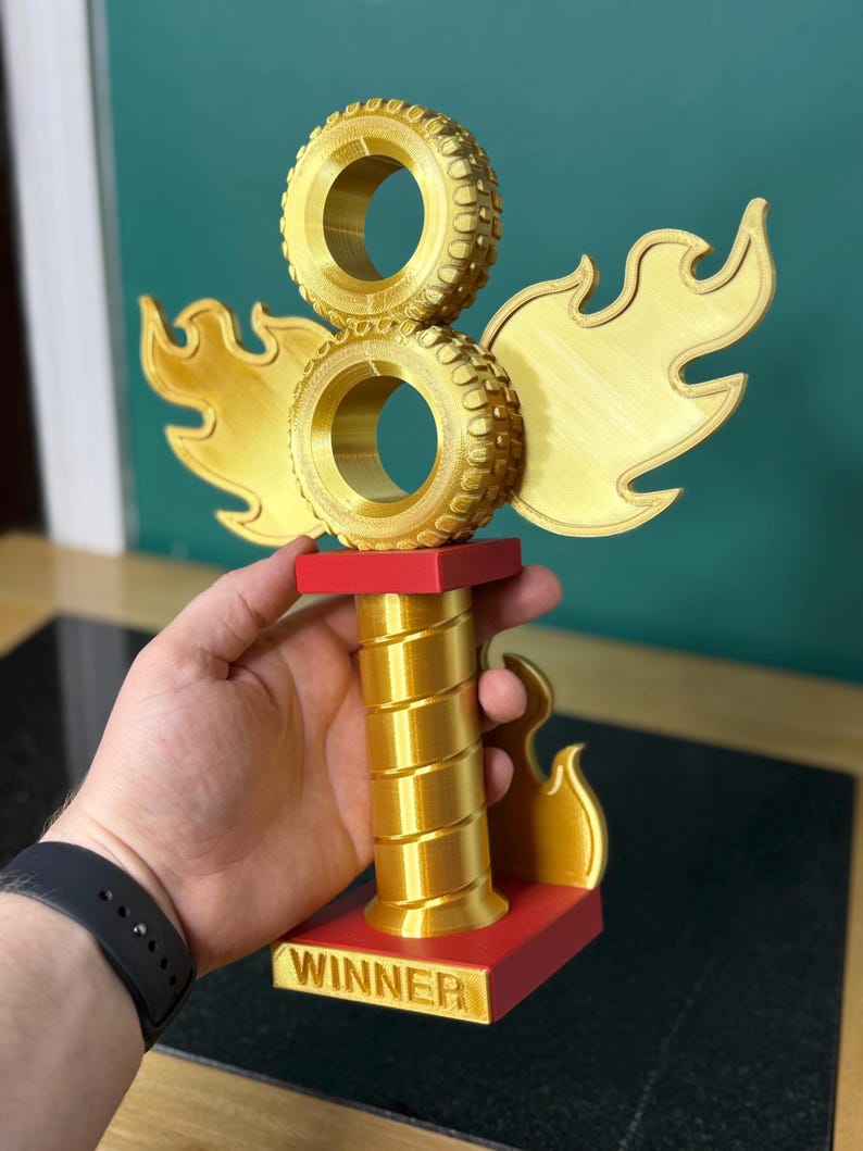 May include: A gold trophy featuring two stacked tyre-shaped elements, flame-shaped wings, and a red base. The trophy has a spiralled column and the word "WINNER" on the base. The trophy is held by a person.