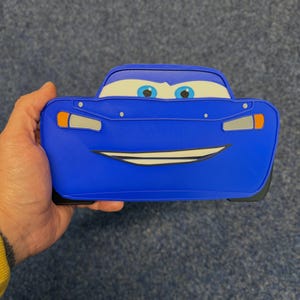 May include: A blue Lightning McQueen-shaped pencil case. The case features the character's face, including eyes, a smiling mouth, and headlights. The case is held in a hand.