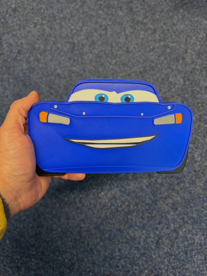 May include: A blue Lightning McQueen-shaped pencil case. The case features the character's face, including eyes, a smiling mouth, and headlights. The case is held in a hand.