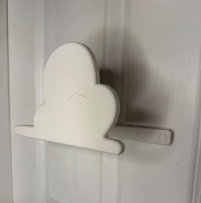May include: A white cloud-shaped wall decor piece, mounted on a white door. The cloud has a rounded shape with a flat base and a small, rectangular extension with two circular holes. The decor is made of a solid, opaque material.