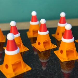 Może przedstawiać: Kolekcja pomarańczowych ozd&oacute;b w kształcie pachołk&oacute;w drogowych, z kt&oacute;rych każda ozdobiona jest miniaturową czerwoną czapką Świętego Mikołaja z białym obszyciem i białym pomponem. Ozdoby ułożone są na ciemnej, odbijającej światło powierzchni.