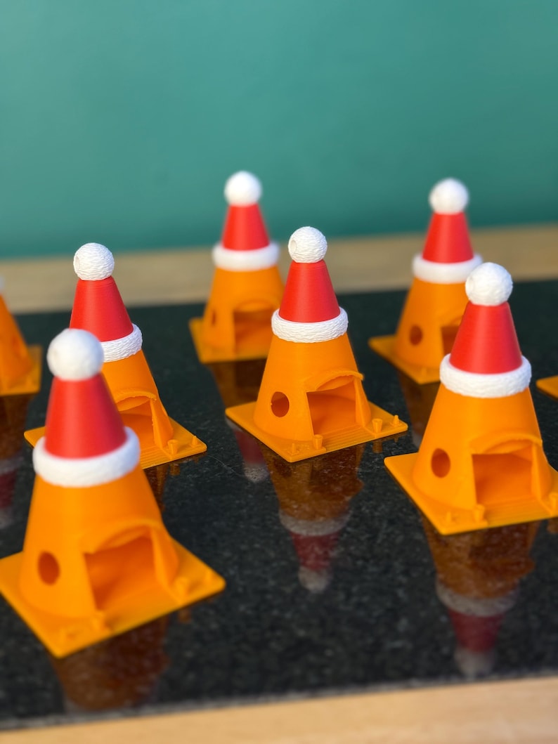 Może przedstawiać: Kolekcja pomarańczowych ozd&oacute;b w kształcie pachołk&oacute;w drogowych, z kt&oacute;rych każda ozdobiona jest miniaturową czerwoną czapką Świętego Mikołaja z białym obszyciem i białym pomponem. Ozdoby ułożone są na ciemnej, odbijającej światło powierzchni.