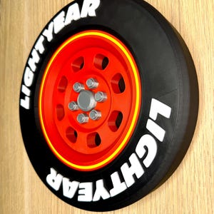 May include: A black and red decorative wheel with the text "LIGHTYEAR" in white. The wheel has a red center with silver-colored bolts and a yellow stripe around the rim. The wheel is mounted on a wooden surface.