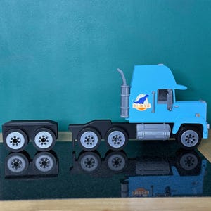 May include: A light blue toy semi-truck with a black trailer. The truck has a silver exhaust pipe and the word "DINOGO" on the side. The trailer has eight wheels, and the truck has six wheels. The toy is on a reflective surface.
