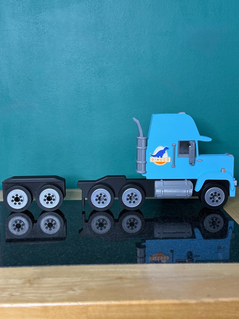 May include: A light blue toy semi-truck with a black trailer. The truck has a silver exhaust pipe and the word "DINOGO" on the side. The trailer has eight wheels, and the truck has six wheels. The toy is on a reflective surface.