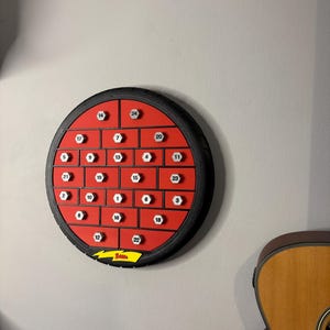 May include: A round, red advent calendar with black trim and numbered compartments. The calendar is mounted on a gray wall. A guitar is partially visible in the background. The calendar has the word "Belisa" and a lightning bolt logo.