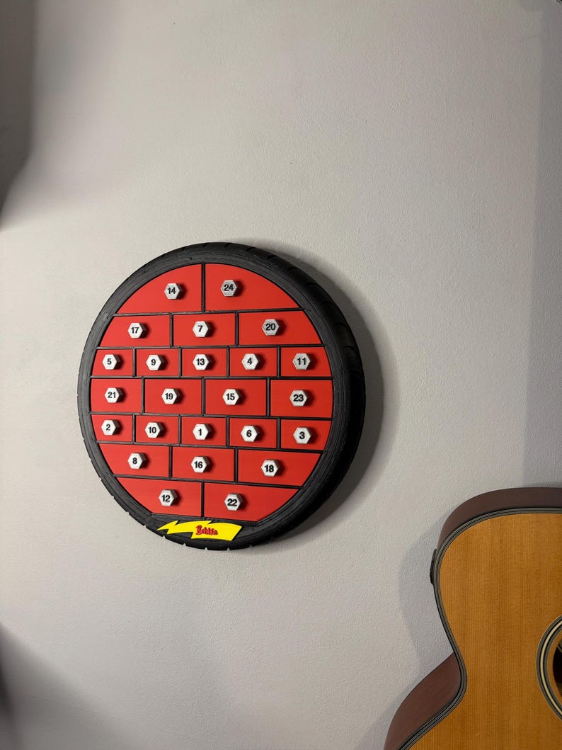 May include: A round, red advent calendar with black trim and numbered compartments. The calendar is mounted on a gray wall. A guitar is partially visible in the background. The calendar has the word "Belisa" and a lightning bolt logo.