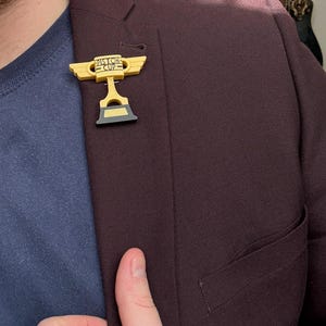 Puede incluir: Un pin de trofeo Piston Cup amarillo y negro est&aacute; fijado a una blazer burdeos oscuro. El pin presenta las palabras "Piston Cup" y un dise&ntilde;o de alas. La blazer se lleva sobre una camisa azul.