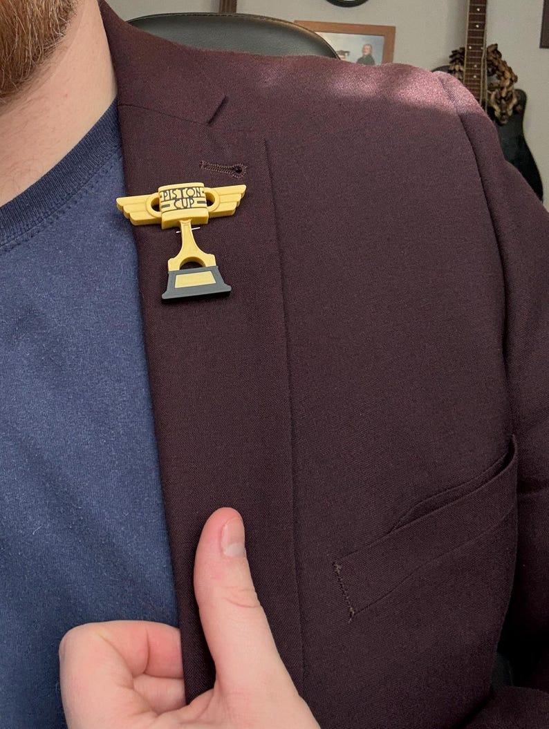 Puede incluir: Un pin de trofeo Piston Cup amarillo y negro est&aacute; fijado a una blazer burdeos oscuro. El pin presenta las palabras "Piston Cup" y un dise&ntilde;o de alas. La blazer se lleva sobre una camisa azul.
