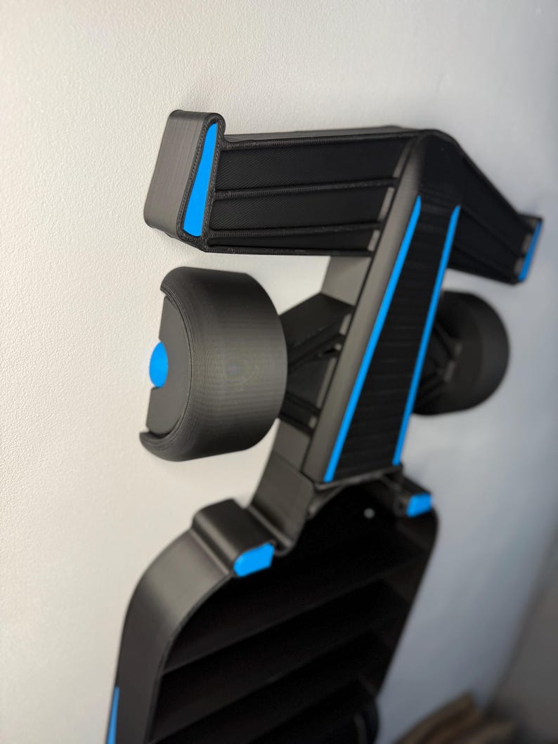 Op de afbeelding: Een zwart en blauw decoratief object, mogelijk een auto-onderdeel, gemonteerd op een witte muur. Het object heeft een complex, geometrisch ontwerp met blauwe accenten en een reeks horizontale lijnen. Het object lijkt 3D-geprint te zijn.