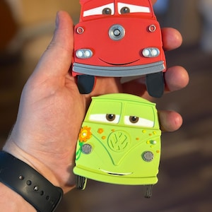 May include: Two toy cars, one red and one green, are held in a hand. The red car has a smiling face and black tires. The green car has a flower and a VW logo. Both cars have cartoon eyes and are designed to look like characters.