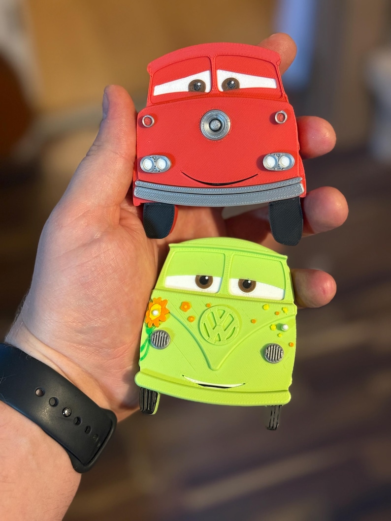 May include: Two toy cars, one red and one green, are held in a hand. The red car has a smiling face and black tires. The green car has a flower and a VW logo. Both cars have cartoon eyes and are designed to look like characters.