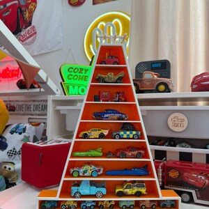 May include: A white and orange pyramid-shaped shelf unit displaying a collection of toy cars. The cars are various colours and designs, including a blue race car and a yellow truck. The shelf is decorated with a Cars theme.