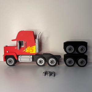 May include: A red toy semi-truck with a yellow '95' lightning bolt graphic on the side. The truck has black wheels and a silver exhaust pipe. The trailer and several silver cylinders are separate.