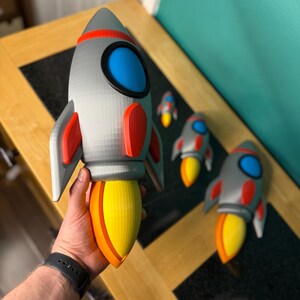 Puede incluir: Un juguete de cohete gris, rojo y amarillo con una ventana azul. El cohete tiene aletas rojas y una llama amarilla y naranja. Hay varios otros cohetes en el fondo.