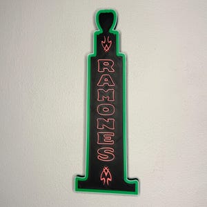 Puede incluir: Un letrero negro y verde con la palabra "RAMONES" en letras rosas. El letrero tiene un borde verde y elementos decorativos en la parte superior e inferior. Est&aacute; montado en una pared blanca.