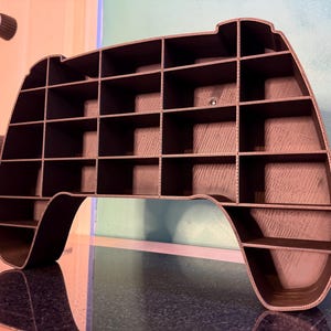 May include: A dark gray, 3D-printed shelf in the shape of a video game controller. The shelf has multiple cubby spaces for storage and display. The controller-shaped shelf is a unique and modern design.