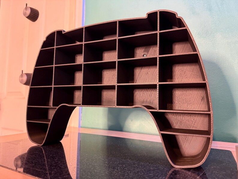 May include: A dark gray, 3D-printed shelf in the shape of a video game controller. The shelf has multiple cubby spaces for storage and display. The controller-shaped shelf is a unique and modern design.