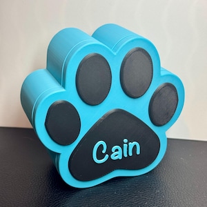 May include: A turquoise blue paw print shaped box with black paw prints and the name "Cain" inscribed on the bottom paw print.