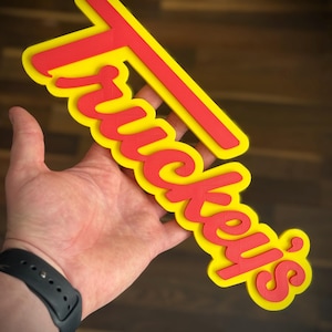 K&ouml;nnte beinhalten: Ein mehrschichtiges Schild in Rot und Gelb mit der Aufschrift "Tuckey's". Das Schild wird in einer Hand gehalten, mit einer schwarzen Uhr am Handgelenk. Die Buchstaben des Schildes sind rot mit gelbem Rand.