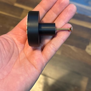May include: A black cabinet knob with a cylindrical body and a round, flat head. The head has a white and gold decorative pattern around the edge. A gold screw extends from the back.