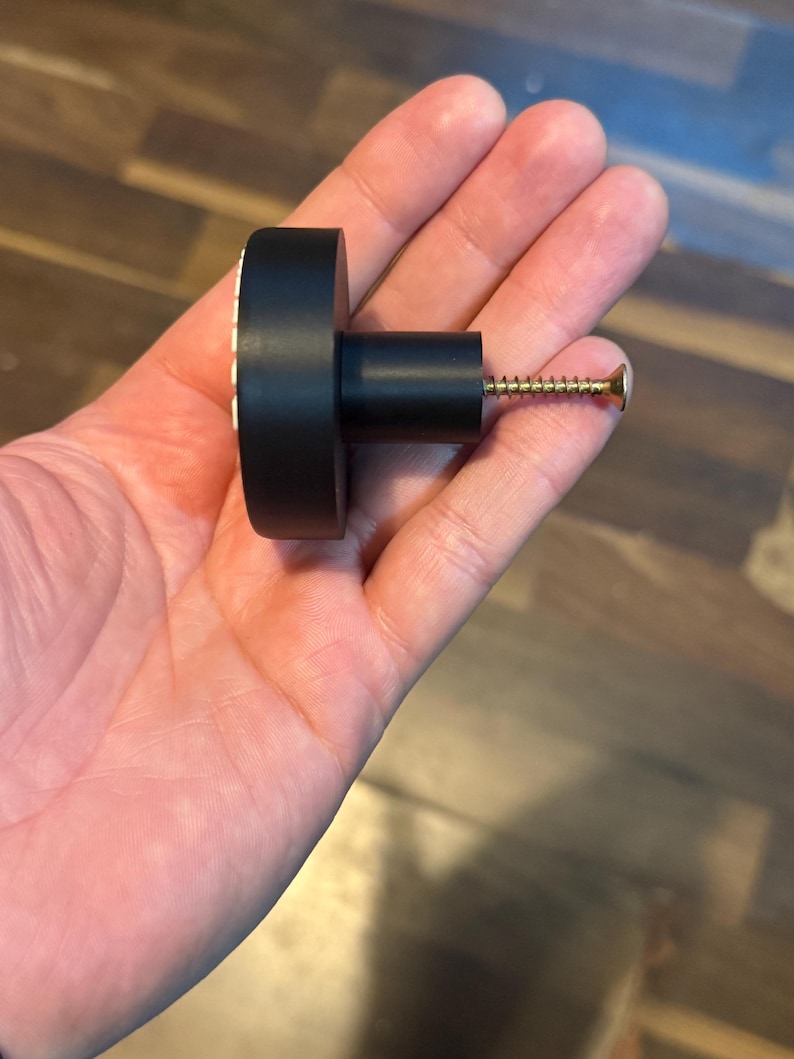 May include: A black cabinet knob with a cylindrical body and a round, flat head. The head has a white and gold decorative pattern around the edge. A gold screw extends from the back.