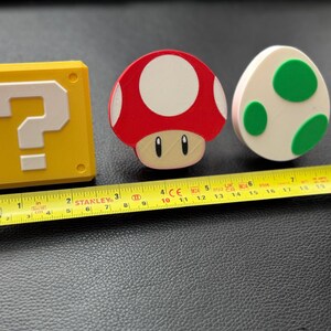 May include: Three colorful, decorative items from the Super Mario Bros. video game series. A yellow question mark block, a red and white mushroom, and a white egg with green spots are displayed. A yellow Stanley tape measure is next to the items.