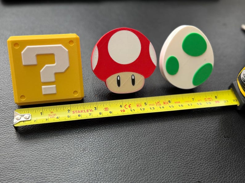 May include: Three colorful, decorative items from the Super Mario Bros. video game series. A yellow question mark block, a red and white mushroom, and a white egg with green spots are displayed. A yellow Stanley tape measure is next to the items.