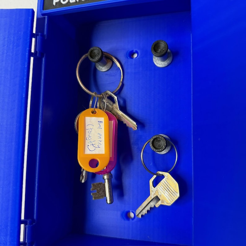 May include: A blue police box-style key holder with keys hanging inside. One key has an orange tag with the text "Bed entry (Tough)". The key holder has a hinged door and metal key rings.