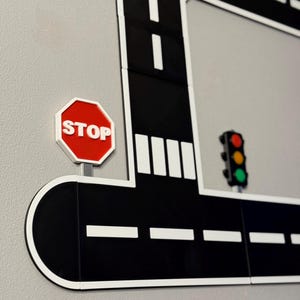 May include: A black and white road sign with a red stop sign and a traffic light. The road has white dashed lines and a white border. The stop sign is red with white text.