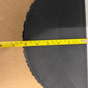 Może przedstawiać: Czarny, p&oacute;łkolisty obiekt o teksturowanej powierzchni jest mierzony ż&oacute;łtą miarką. Miarka pokazuje długość około 23 cm. Tło stanowi jasnobrązowa powierzchnia i biała powierzchnia.