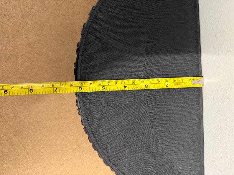 Może przedstawiać: Czarny, p&oacute;łkolisty obiekt o teksturowanej powierzchni jest mierzony ż&oacute;łtą miarką. Miarka pokazuje długość około 23 cm. Tło stanowi jasnobrązowa powierzchnia i biała powierzchnia.