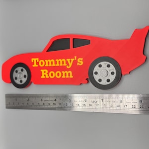 May include: A red car-shaped sign with the text "Tommy's Room" in yellow. The sign has black wheels and windows. A ruler is placed below the sign, measuring up to 24 inches.