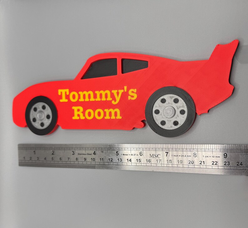 May include: A red car-shaped sign with the text "Tommy's Room" in yellow. The sign has black wheels and windows. A ruler is placed below the sign, measuring up to 24 inches.
