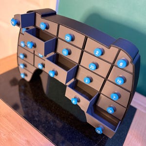 May include: A gray and blue advent calendar with 24 numbered drawers. Each drawer has a blue knob. The calendar is shaped like a rounded rectangle and sits on a dark surface.
